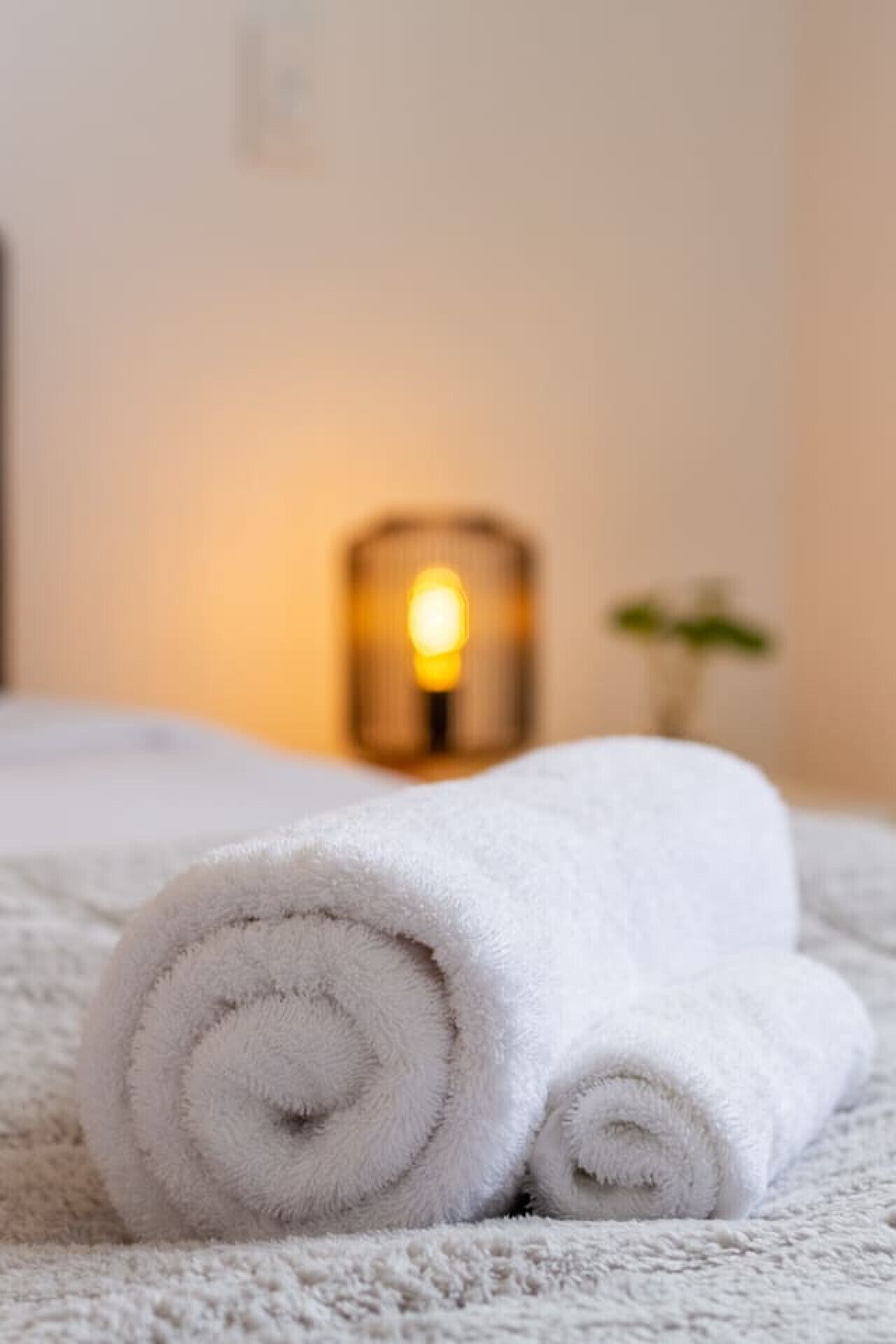 A neatly rolled white towel on a textured white bedspread with a soft glow from a candle in the background.
