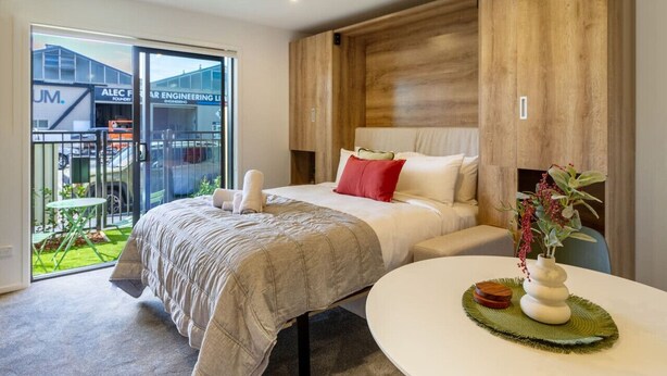 A modern hotel room with a large bed, a round table, and a view of an outdoor area with a building labeled "Alec Farber Engineering Ltd." - Cozy Studio Escape in Wilmer (Christchurch)