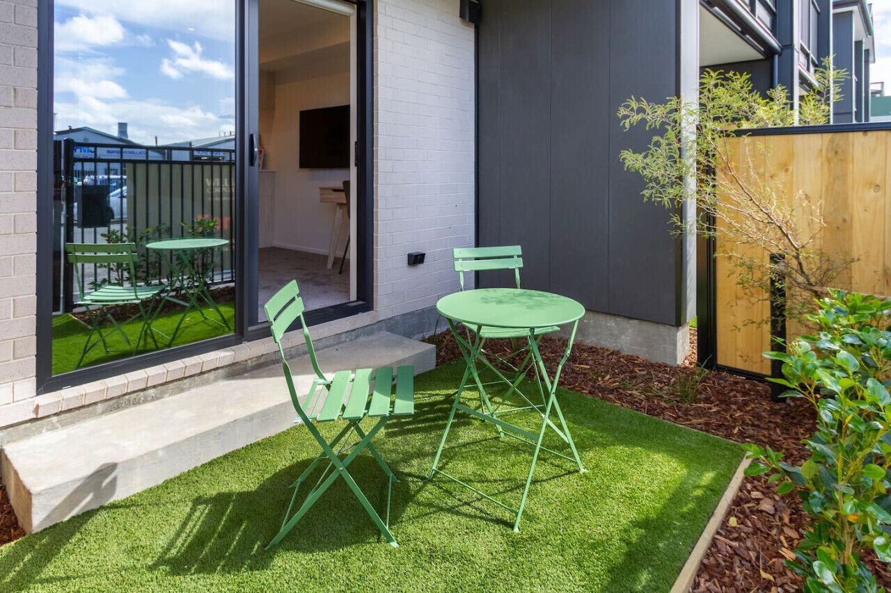 A patio with green chairs and a table, a small garden area with artificial grass, and a wooden fence.