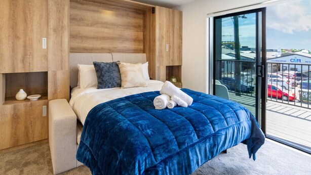 A modern bedroom with a large bed, wooden headboard, and a view of a parking area through a glass door.