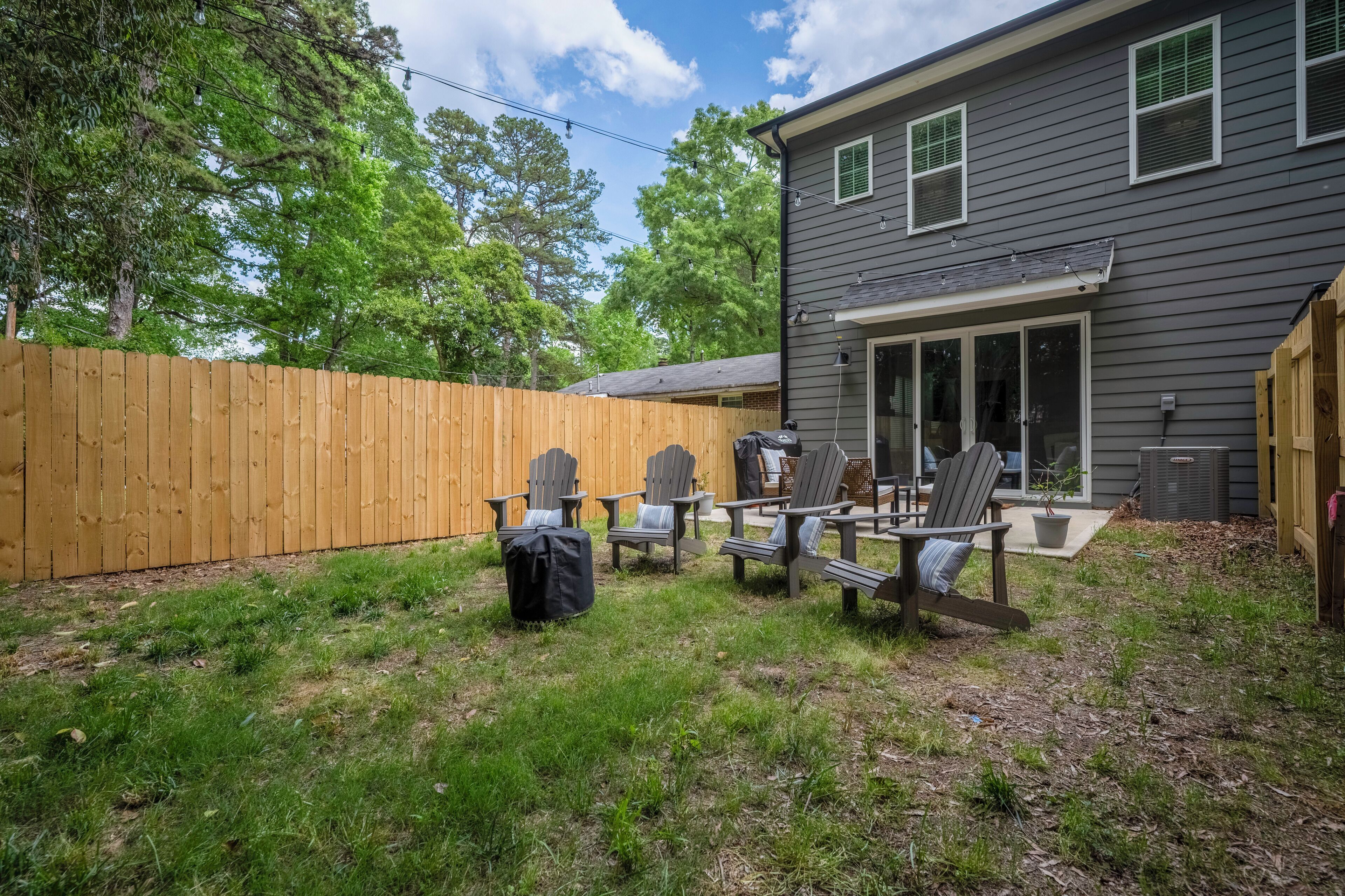 Un patio trasero con cerca de madera, mobiliario exterior con sillas Adirondack, y una casa de fachada gris con un porche cubierto.