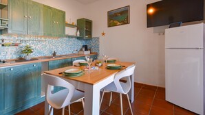 Une cuisine avec un mur recouvert de carreaux, une table à manger en bois avec des chaises blanches, un réfrigérateur blanc et un petit îlot central doté d’un évier et d’un plan de travail.
