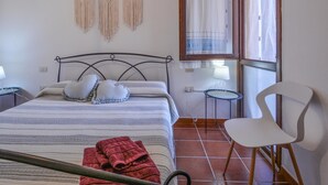 Une chambre à coucher avec un lit, deux oreillers, une serviette rouge, une chaise blanche, une petite table avec une lampe et des décorations murales en macramé.