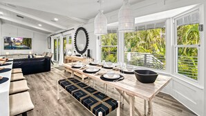 A modern kitchen with a long dining table set for six, a black leather sofa, and large windows with a view of greenery.