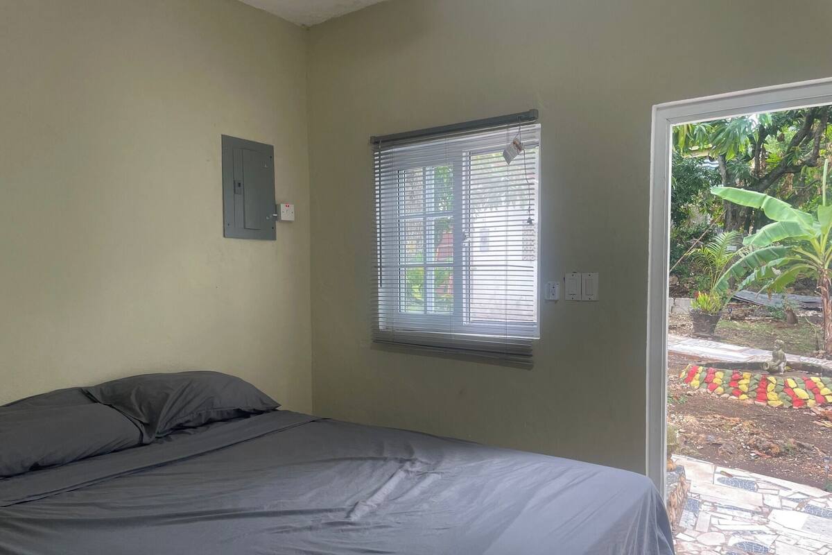 Ein Schlafzimmer mit Bett, einem Fenster mit Jalousien, einem kleinen Stromkasten und Blick auf einen Garten.