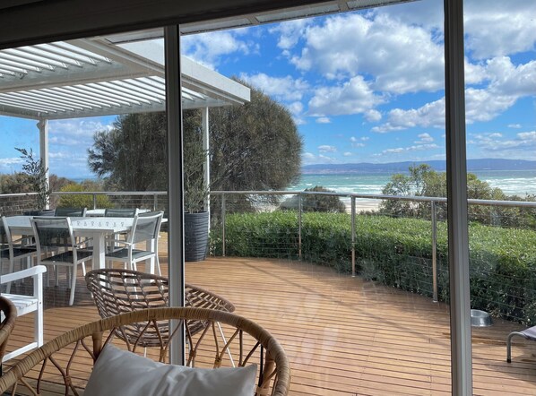 Un espace extérieur abrité, avec une table et des chaises, une plante en pot, et une vue sur la mer et les montagnes.