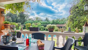Une terrasse extérieure avec une table dressée pour le repas, entourée de chaises, et donnant sur un jardin où l’on peut admirer diverses plantes et une piscine.