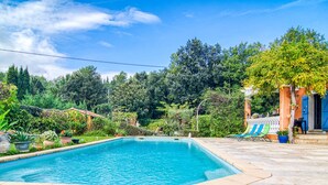 Une piscine rectangulaire, bordée d’une terrasse carrelée et entourée d’une végétation luxuriante et d’arbres.