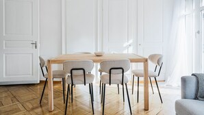 Ein Essbereich mit Holztisch und sechs Stühlen, weißen Wänden und einem großen Fenster mit durchsichtigen Vorhängen.