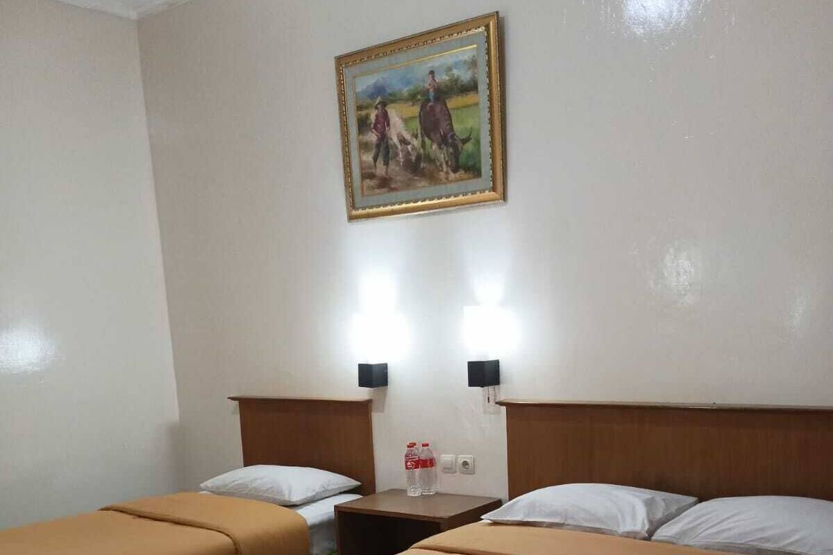 A hotel room with two beds, each with a green skirt and a brown bedspread. There is a nightstand with two bottles of water and a small lamp. A framed painting hangs on the wall above the beds.