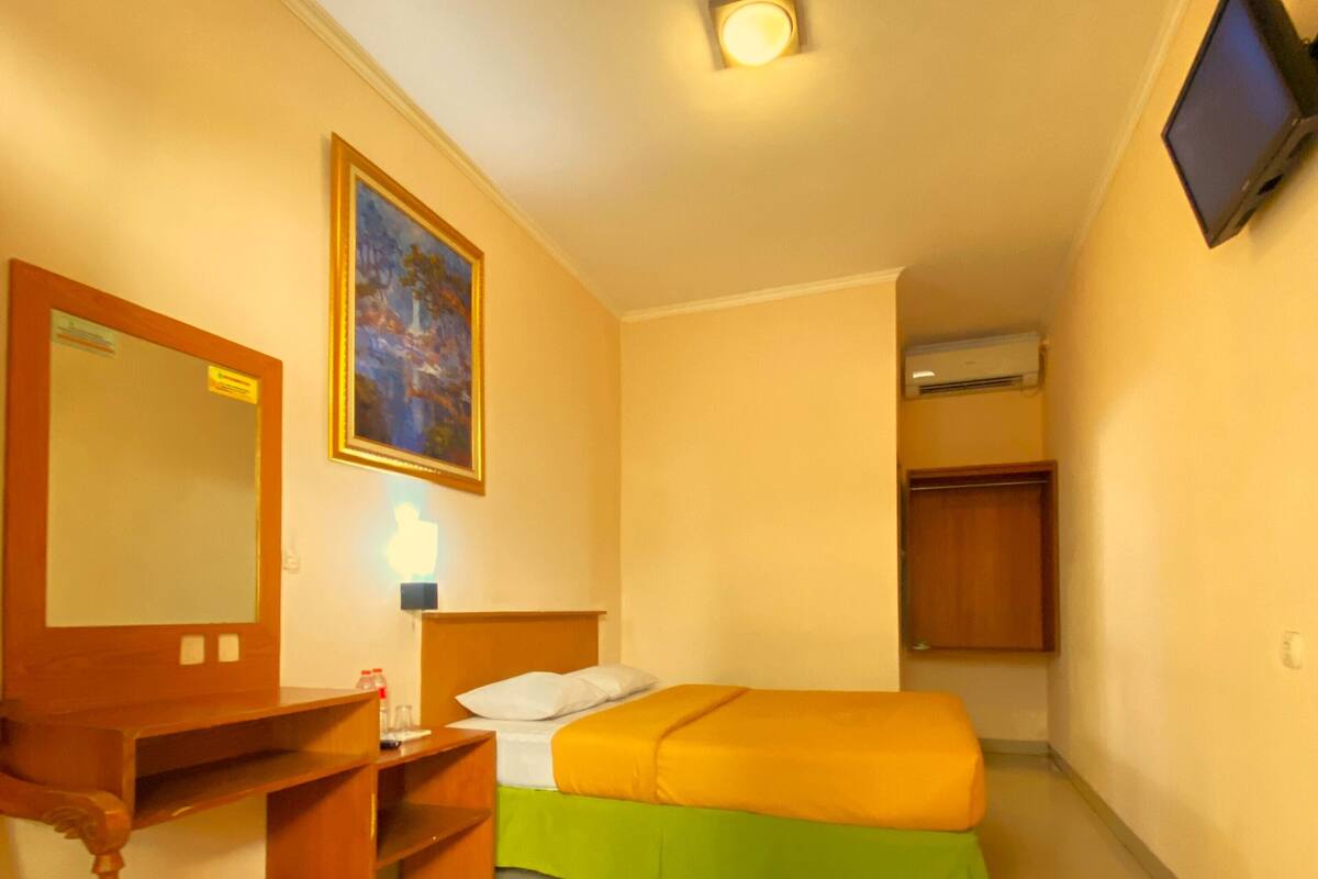 A hotel room with a bed, a desk with a chair, a mirror, a painting, a television, and an air conditioning unit.