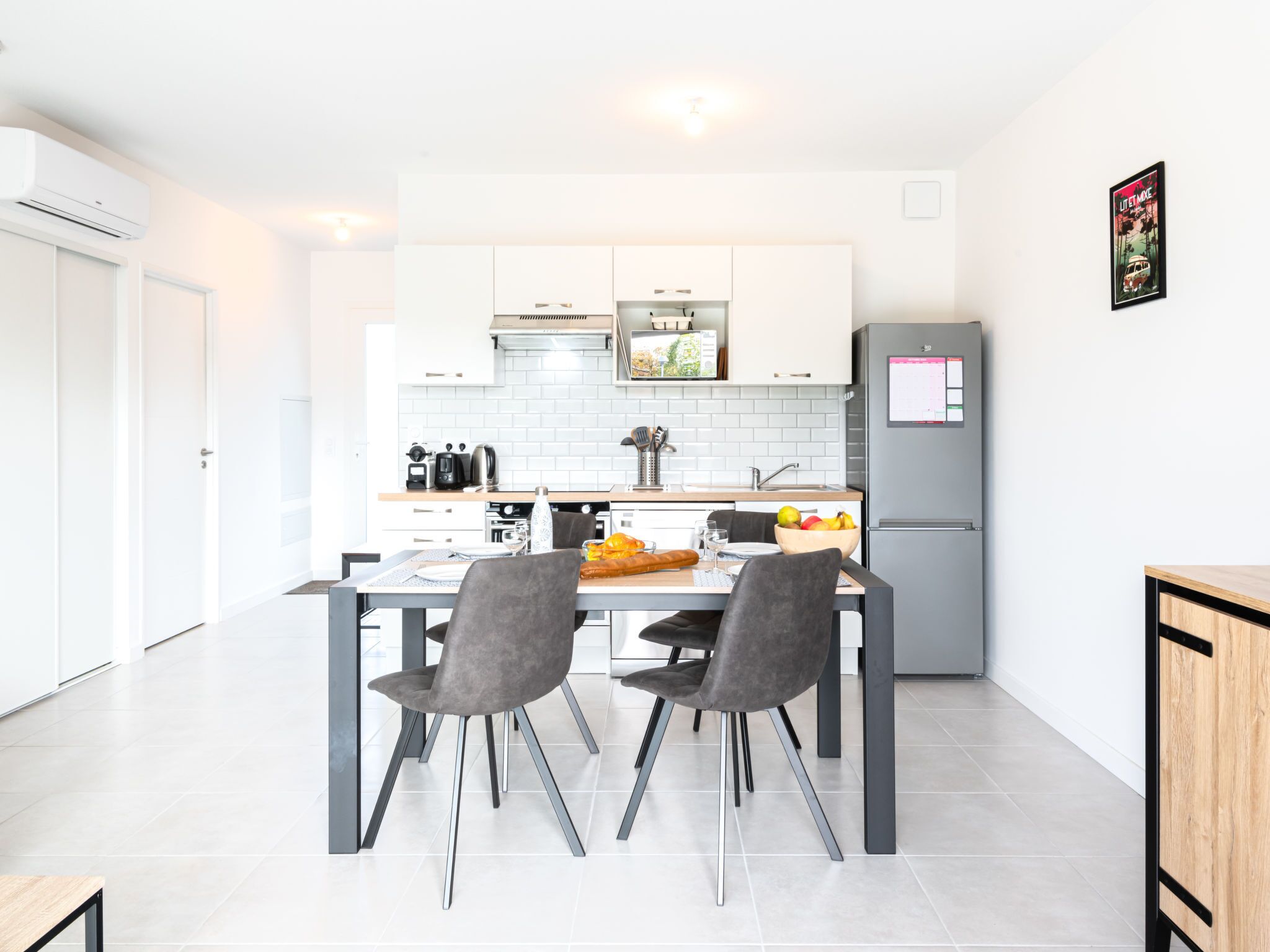 Una cocina moderna con armarios blancos, un refrigerador gris y un comedor con dos sillas grises y una mesa preparada para dos.