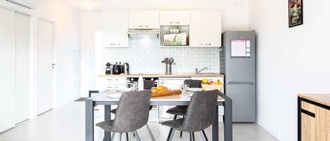 Una cocina moderna con armarios blancos, un refrigerador gris y un comedor con dos sillas grises y una mesa preparada para dos.