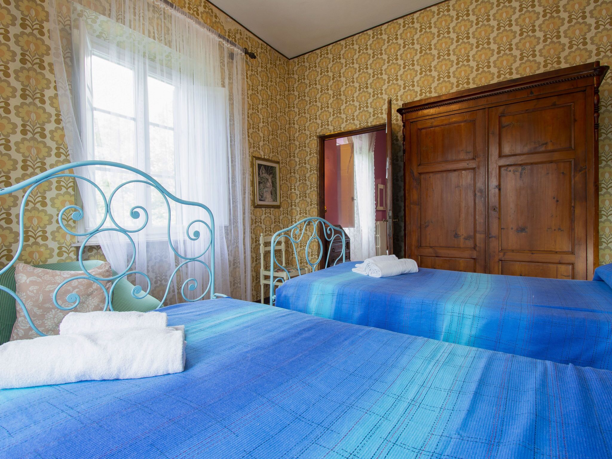 A bedroom with two beds, a wooden wardrobe, and a window with sheer curtains.