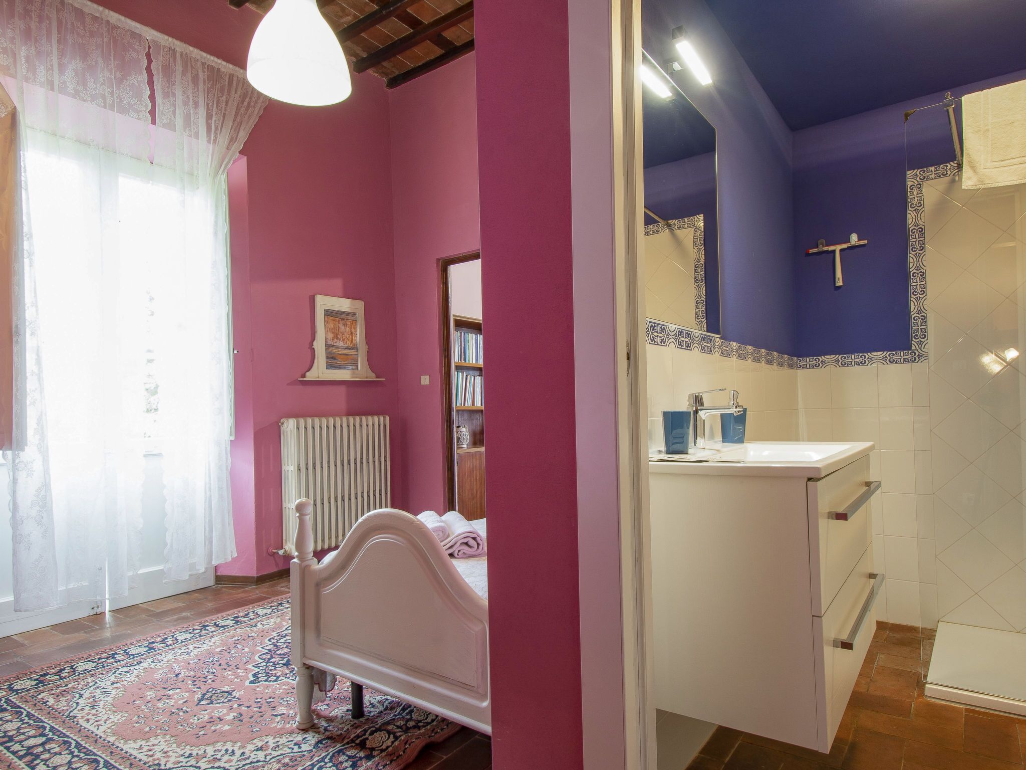 A bedroom with a bed, a radiator, a bookshelf, and a bathroom with a sink and mirror.