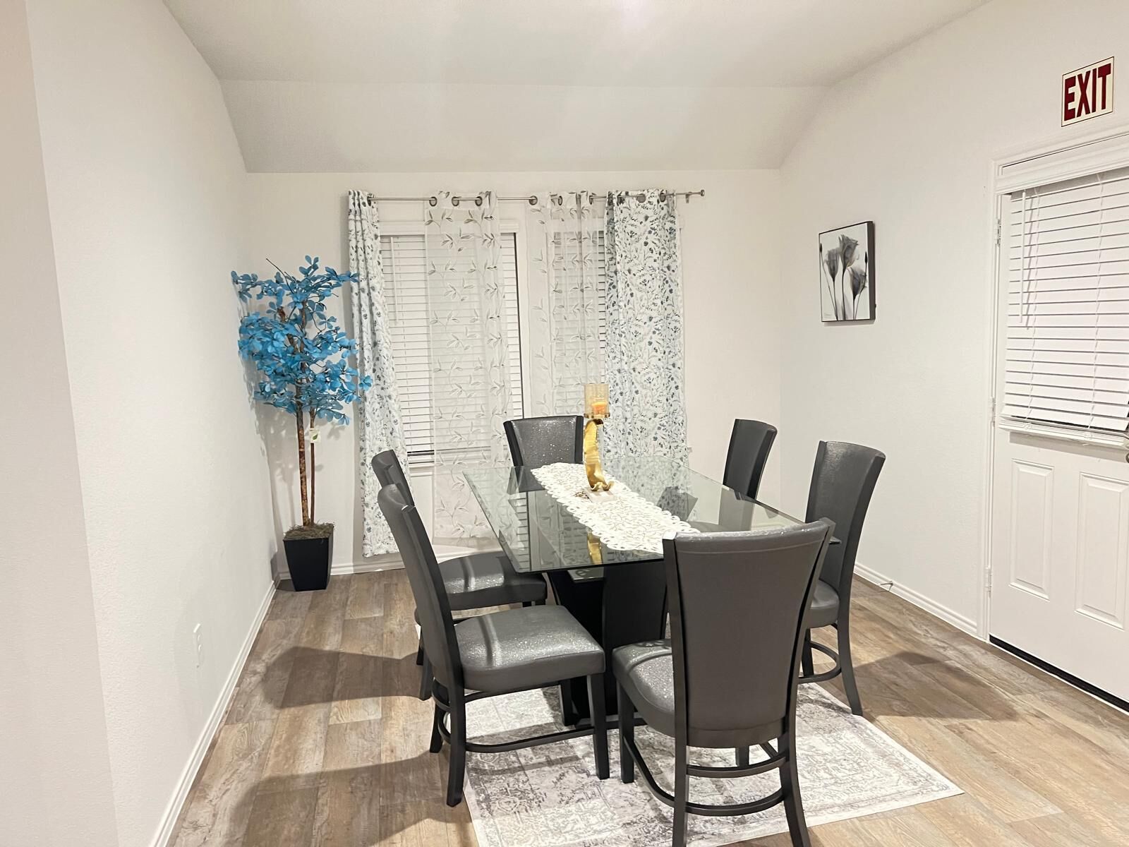 A dining area with a glass table, grey chairs, a potted plant, and a patterned rug.