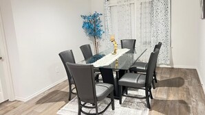 Een eetgedeelte met een glazen tafel, zes stoelen, een decoratieve blauwe plant en kamergordijnen met een patroon.