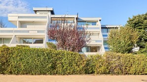 Ein modernes, mehrgeschossiges Gebäude mit flachem Dach, großen Fenstern und einer gepflegten Hecke im Vordergrund.