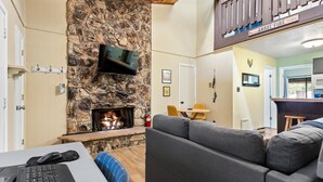 Een gezellige woonkamer met een stenen open haard, een flatscreen televisie erboven, een grijze bank en een houten plafond.
