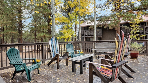 Een houten terras met Adirondack-stoelen en een tafel, omgeven door bomen en een gebouw met een balkon.