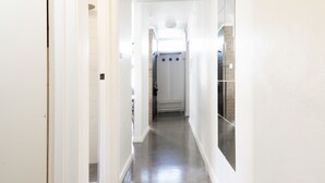 Un pasillo estrecho con paredes blancas y suelo de baldosas, que conduce a un baño con zona de ducha.