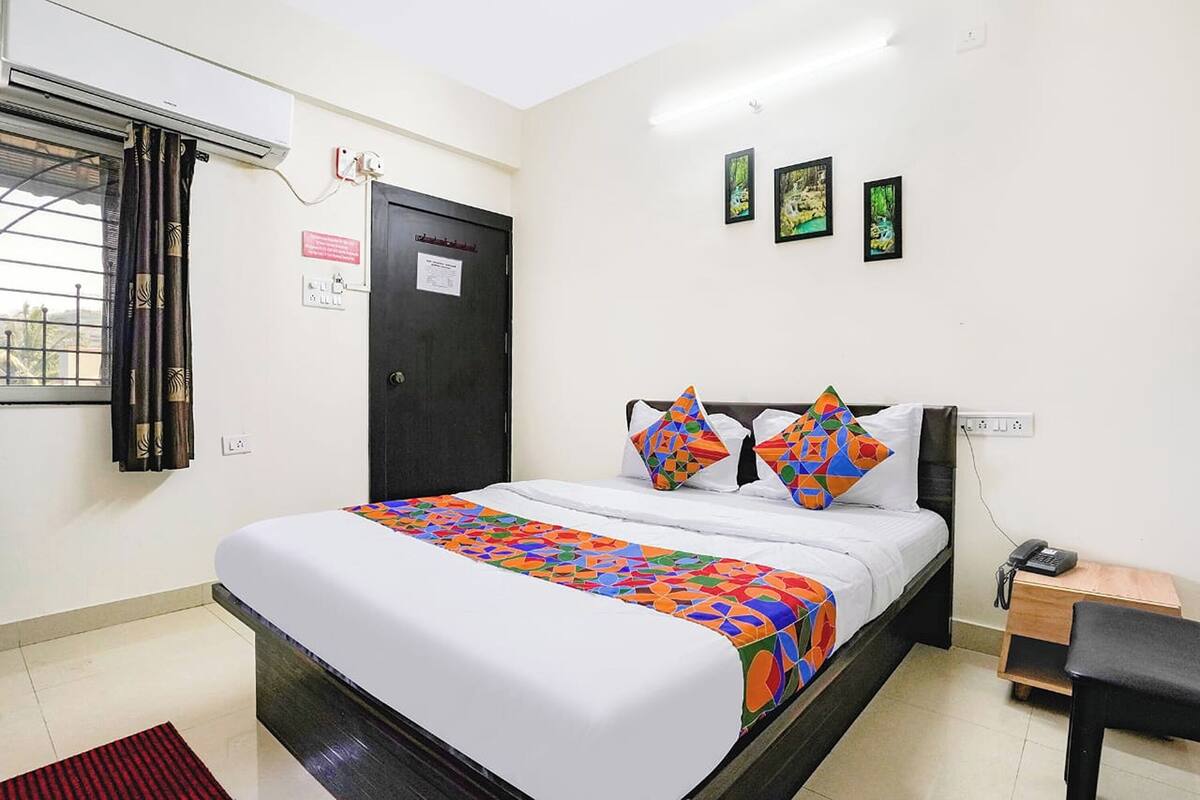 A hotel room with a bed, a window with curtains, a door, and a small table with a phone.
