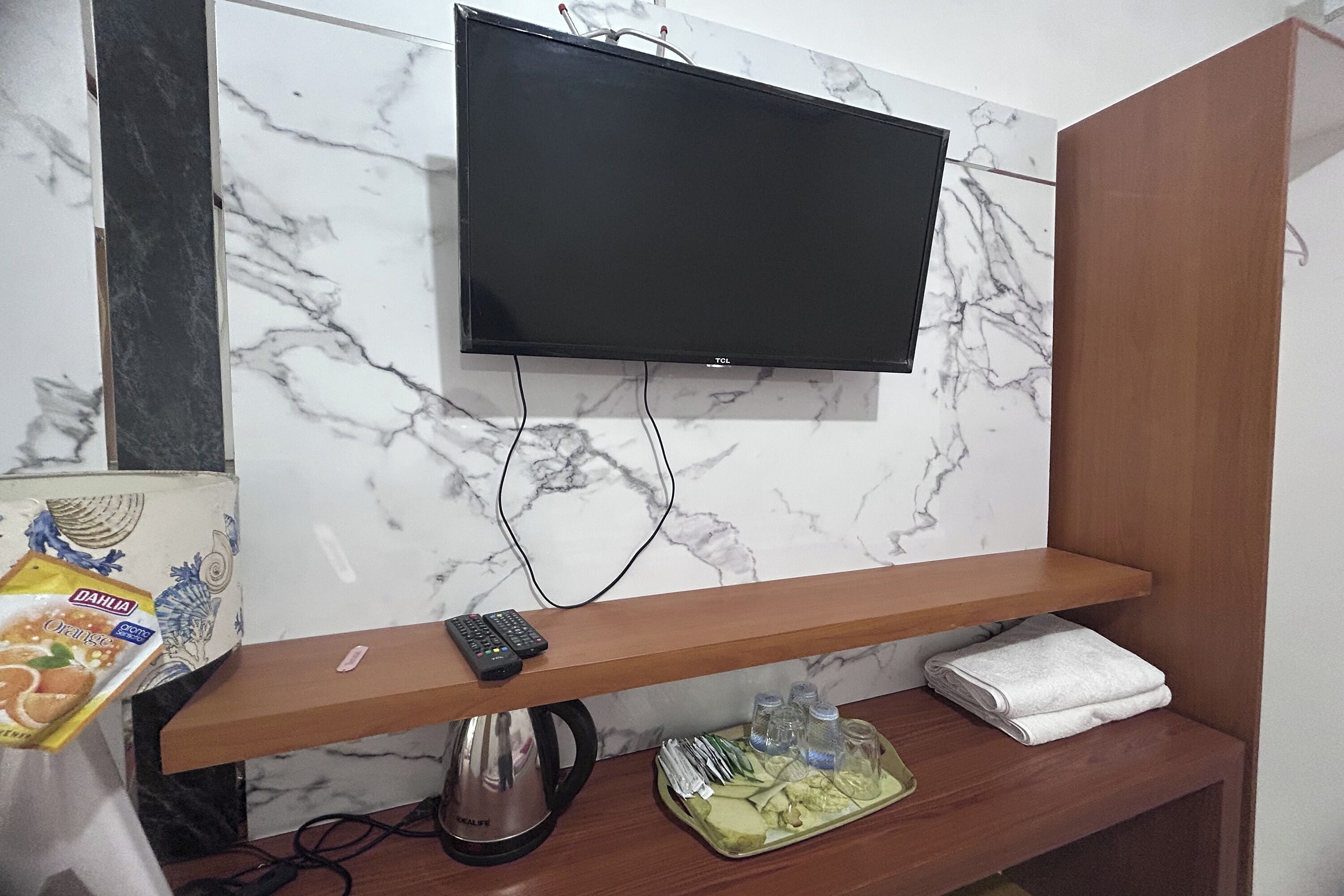 Un televisor de pantalla plana montado en una pared de mármol, un estante de madera debajo, un hervidor de agua, una bandeja con comida y una caja de aperitivos.