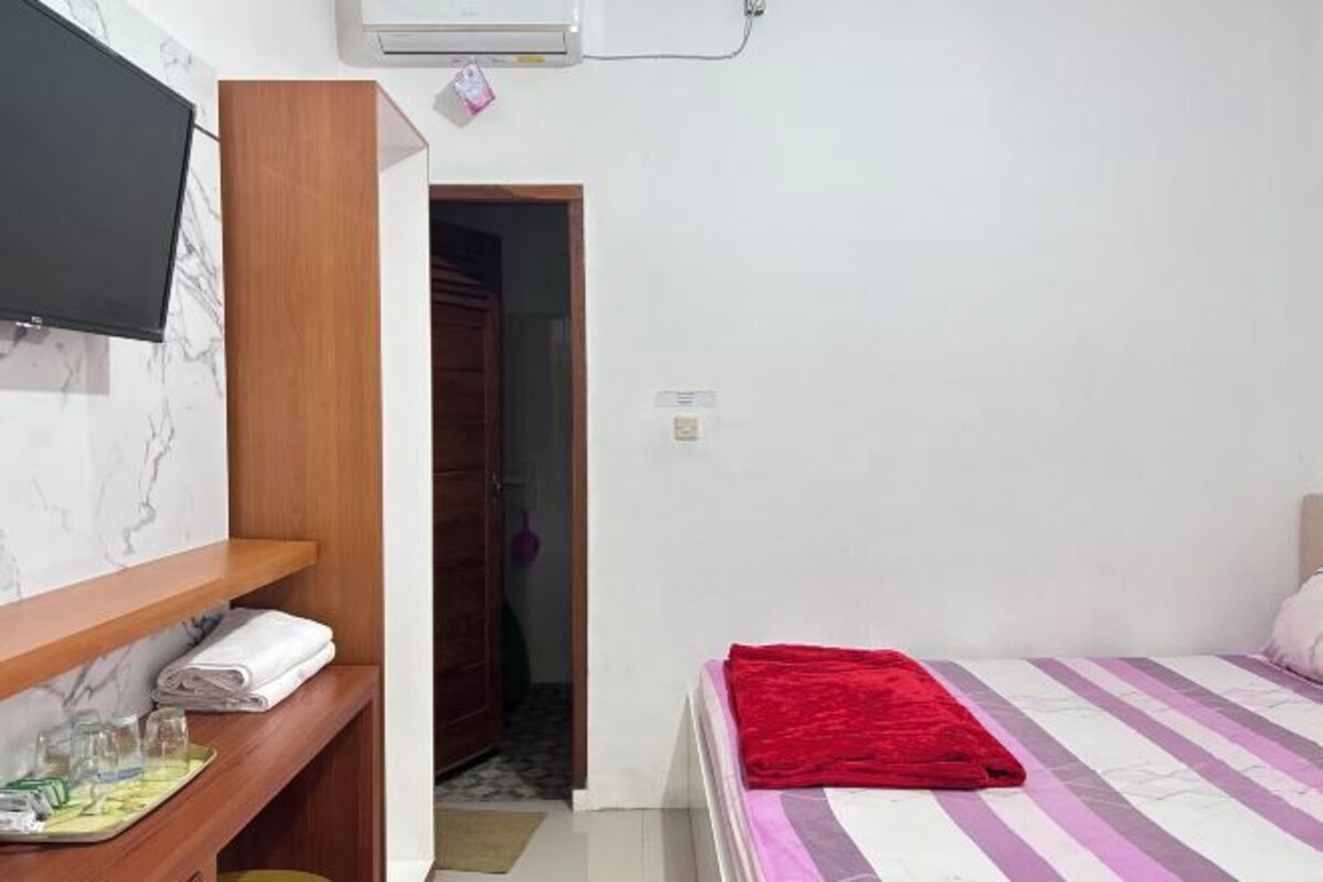 A bedroom with a bed, a TV mounted on a wall, a wooden shelf with towels, and a door leading to another room.