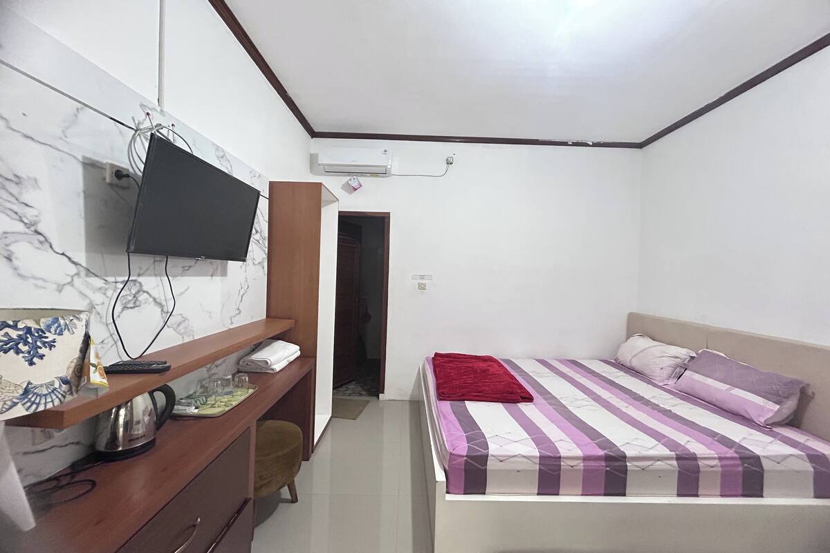 Ein Schlafzimmer mit einem Bett, einem Schreibtisch mit Computer, einem an der Wand befestigten Fernseher und einer Tür zu einem anderen Raum.
