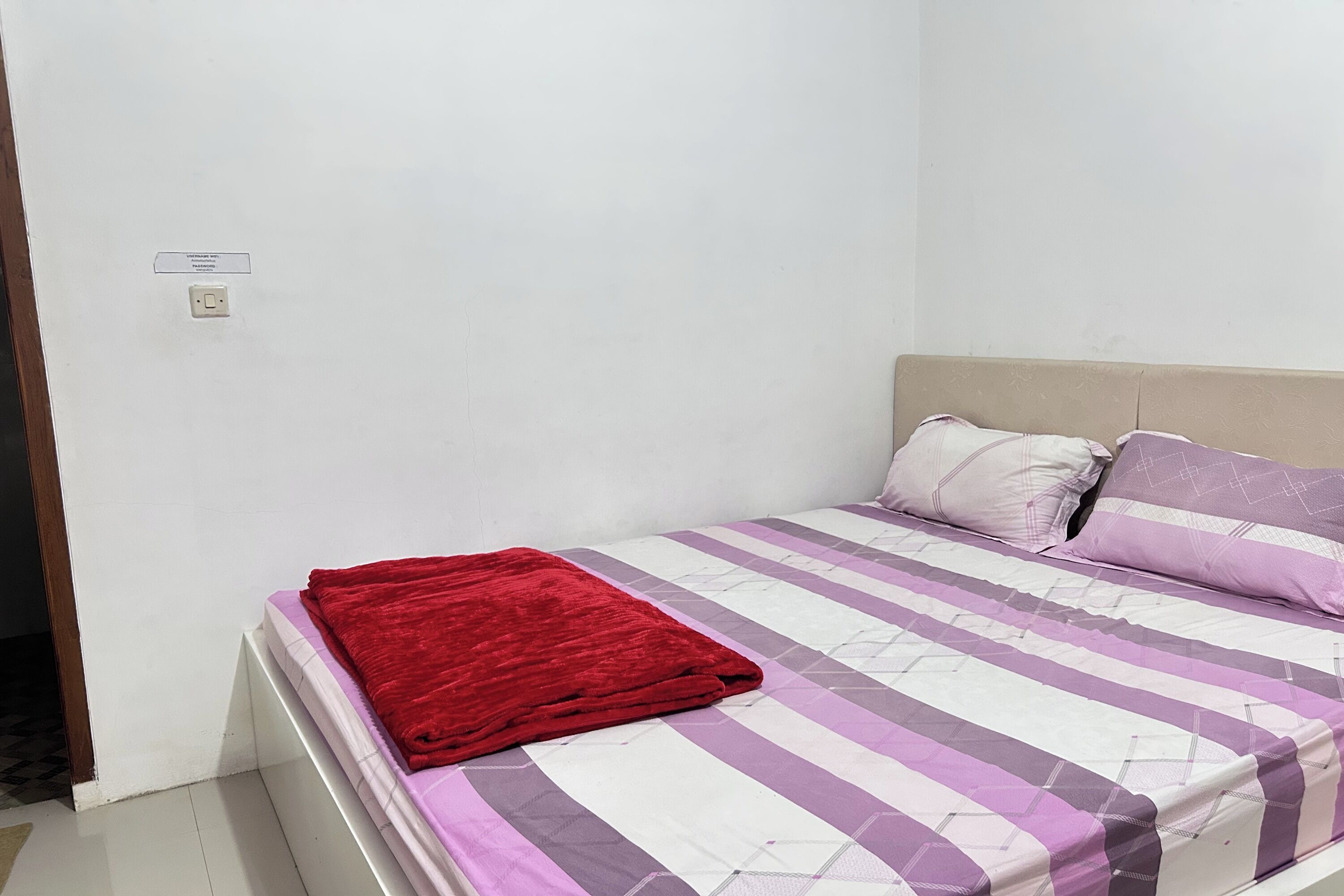 Een eenpersoonskamer met een hoofdeinde, een rode deken en een sprei met paarse en witte strepen.