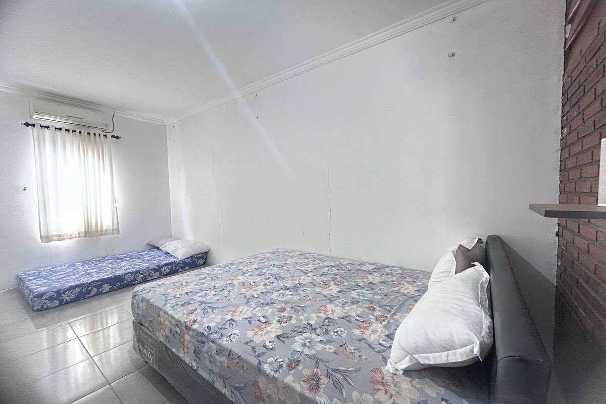 Ein Schlafzimmer mit einem großen Bett, einem kleineren Bett, einem Fenster mit Vorhängen und einer Ziegelwand.