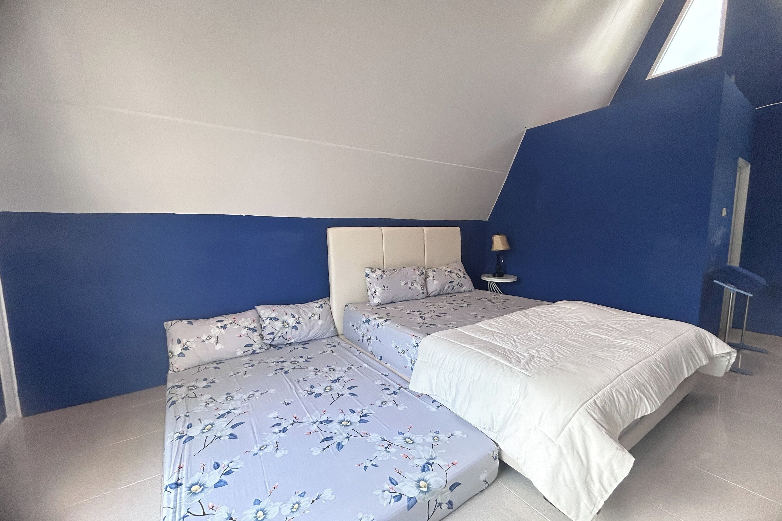 Ein Schlafzimmer mit schrägen Wänden, zwei Betten mit Blumen- und schlichten Bettbezügen, einem Nachttisch mit Lampe und Blick auf den Himmel durch ein Dachfenster.
