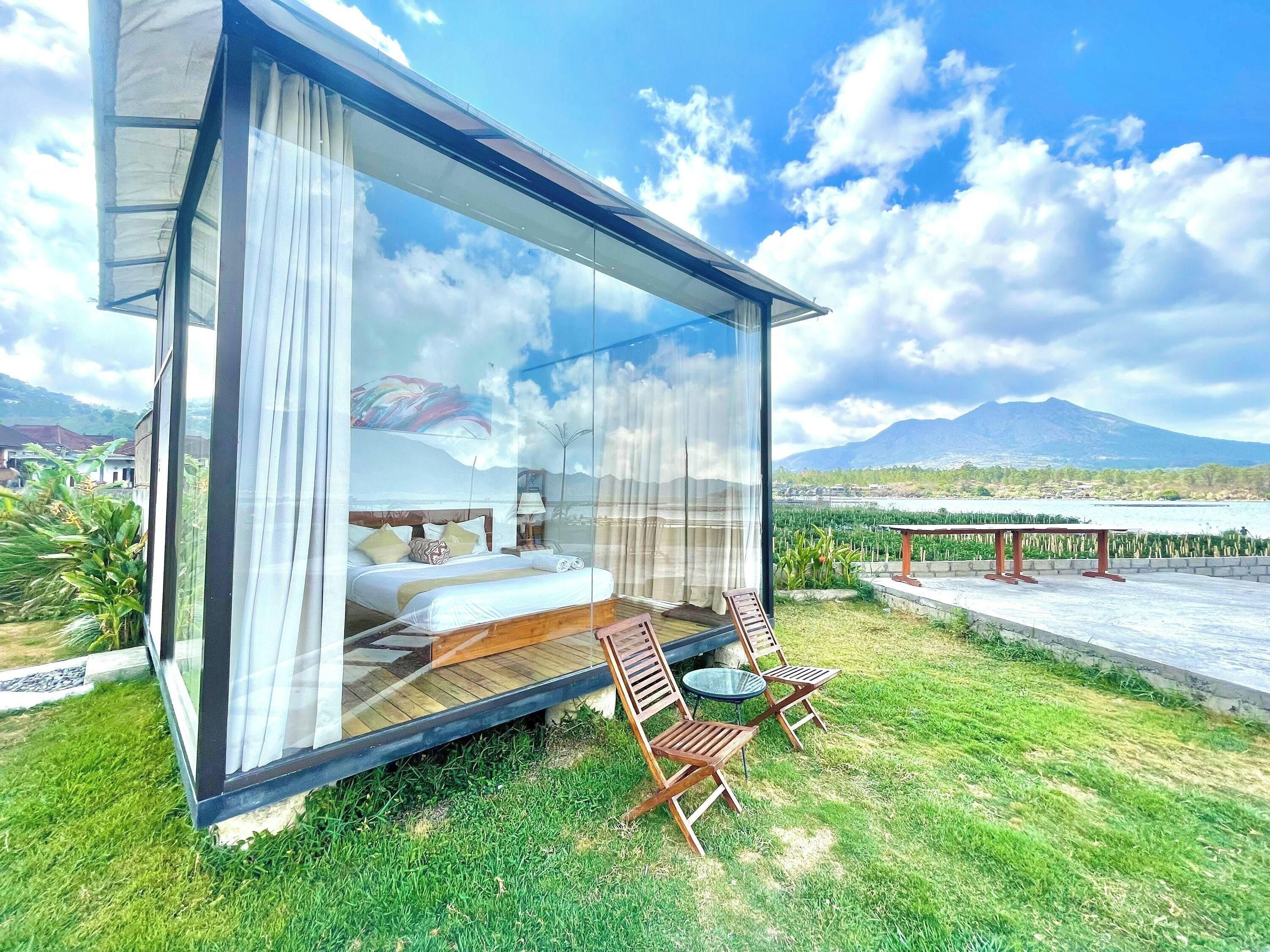 A modern glass room with a bed, curtains, and a view of a mountain and water.