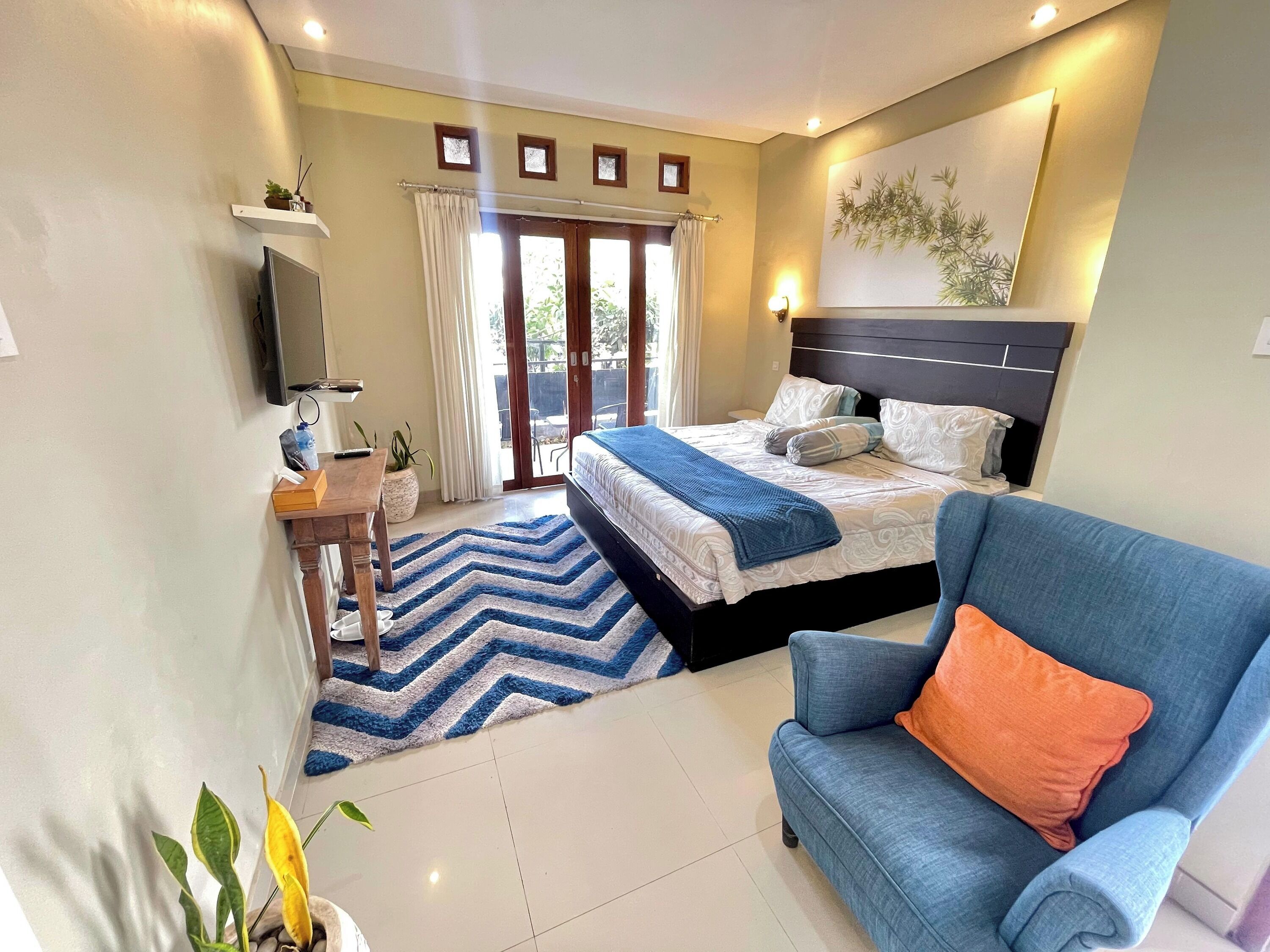 Un dormitorio con una cama, un sillón azul, un escritorio con una planta y un televisor en la pared.