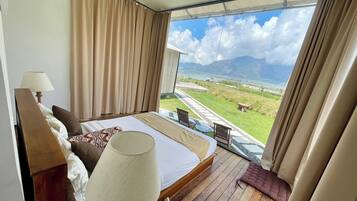 A bedroom with a large bed, a bedside table, a lamp, and a view of a grassy area and mountains through a large window.