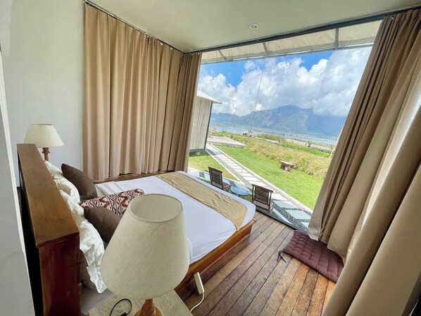 Un dormitorio con una cama grande, una mesita de noche, una lámpara y vistas a un área verde y montañas a través de un ventanal amplio.
