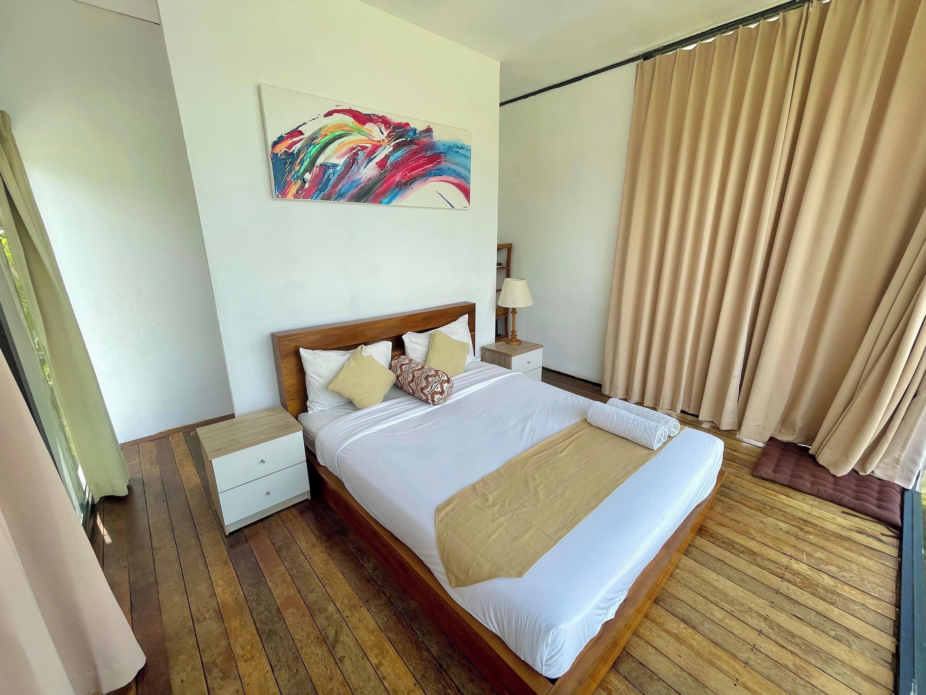 A hotel room with a wooden bed, white bedding, a nightstand with a lamp, a painting on the wall, and a window with curtains.