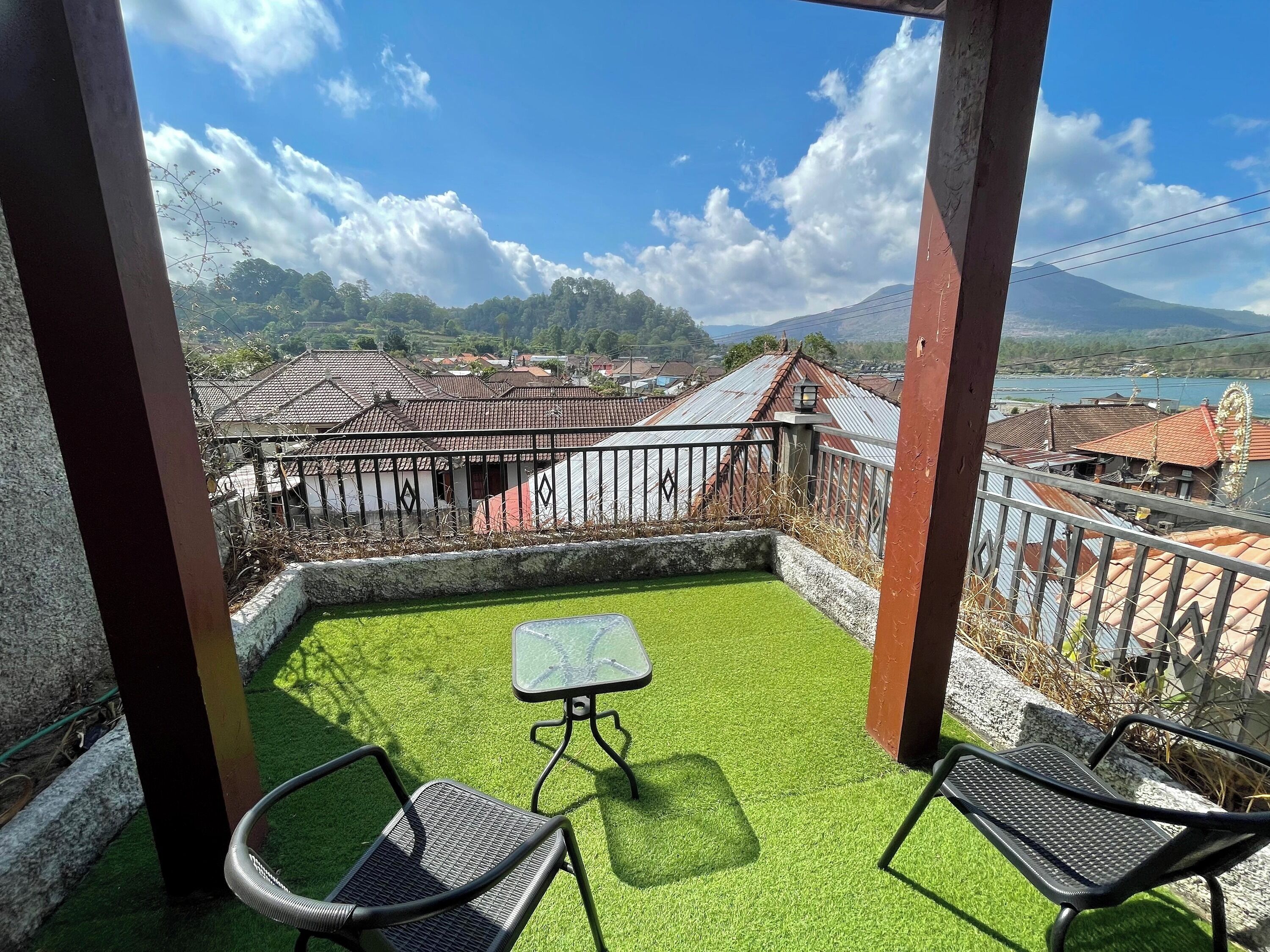 Un balcón con césped artificial, dos sillas de metal y una pequeña mesa. Se observa una zona residencial con casas y una montaña al fondo.
