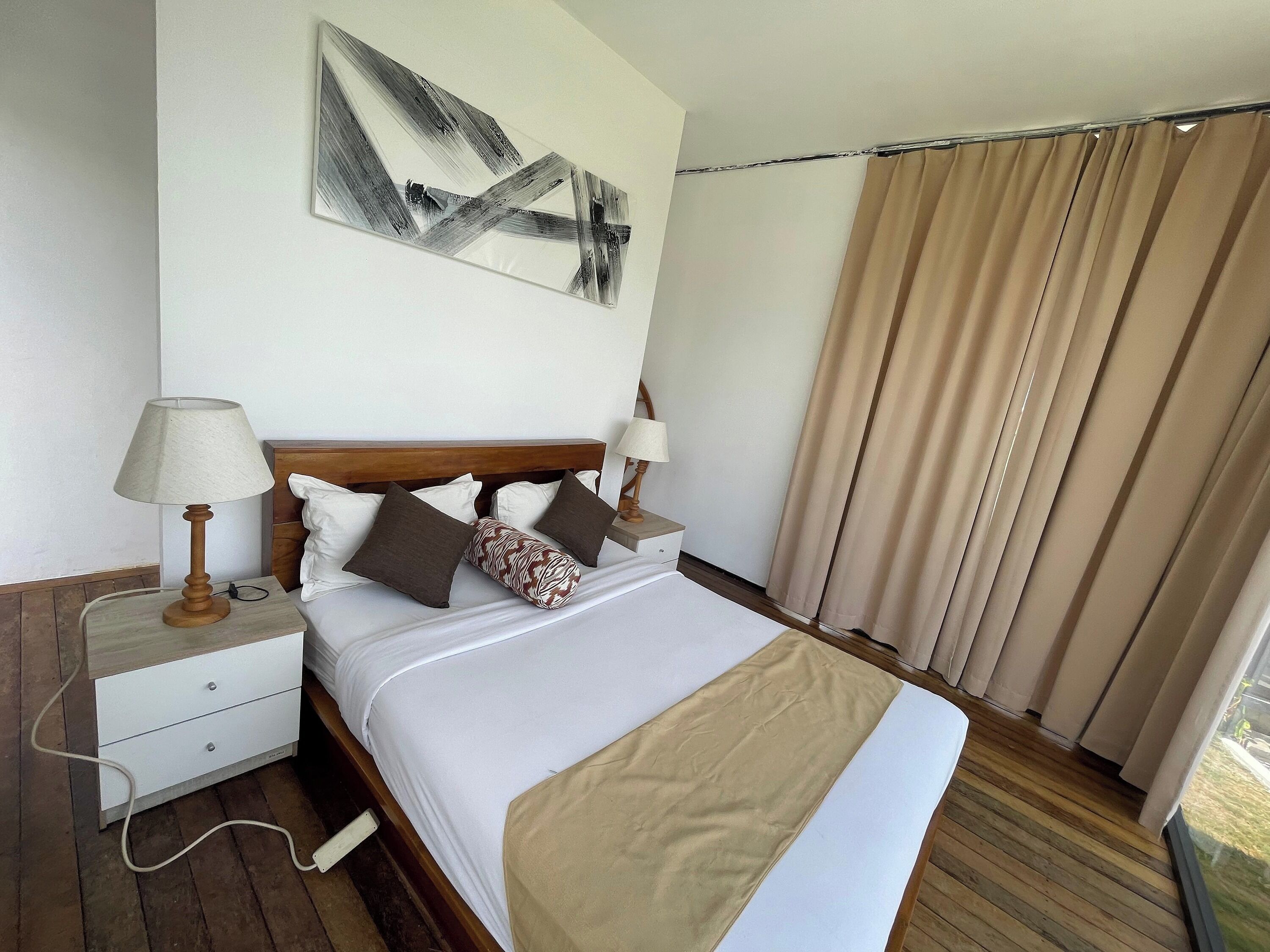 A hotel room with a wooden bed, white bedding, a beige throw blanket, a nightstand with a lamp, and a large window with curtains.