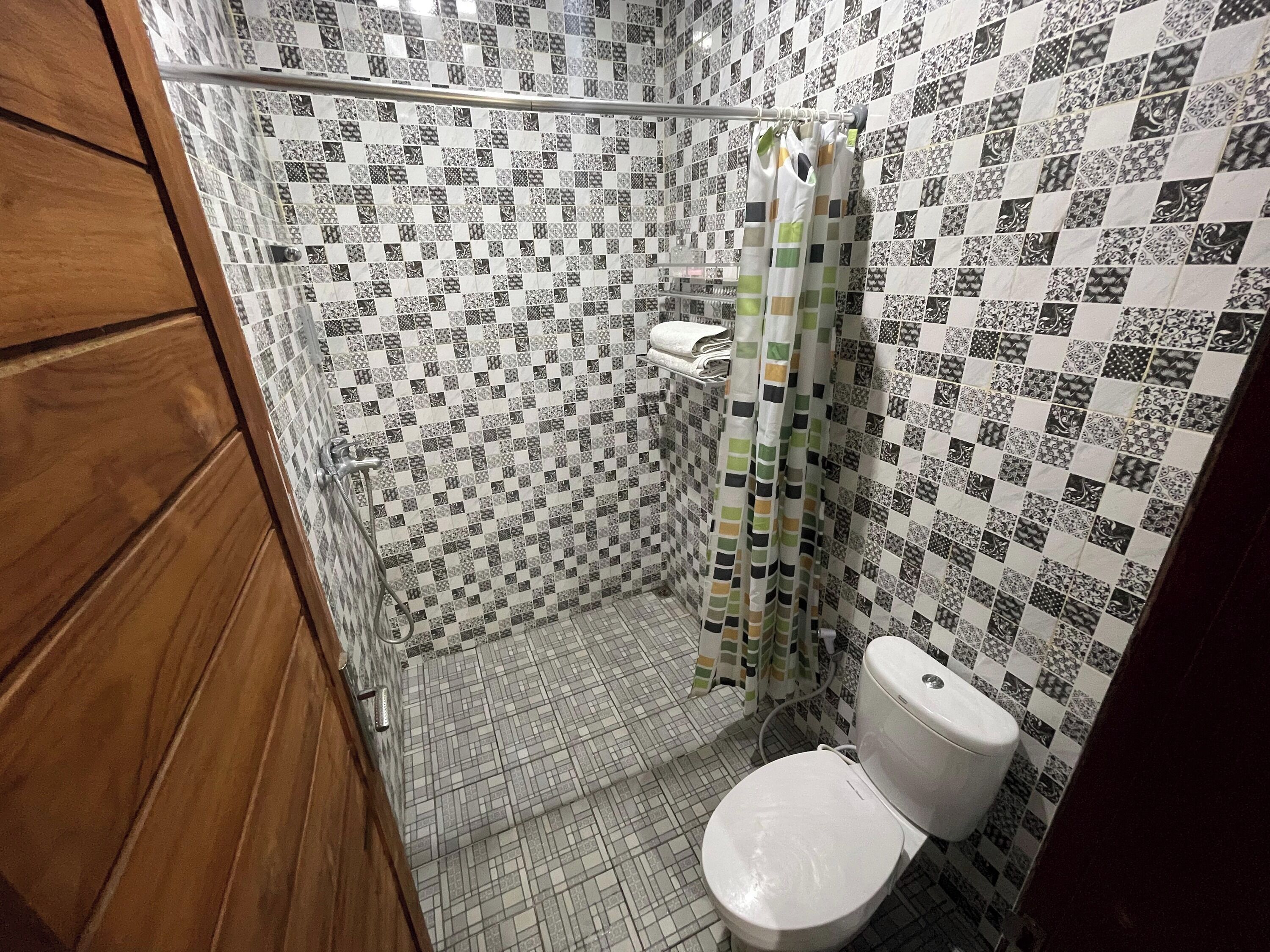 Une salle de bain avec un carrelage au sol, des toilettes, une douche avec un rideau à motifs et un meuble en bois.