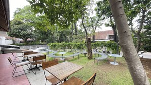 An outdoor seating area with wooden tables and chairs, surrounded by trees and a grassy lawn.