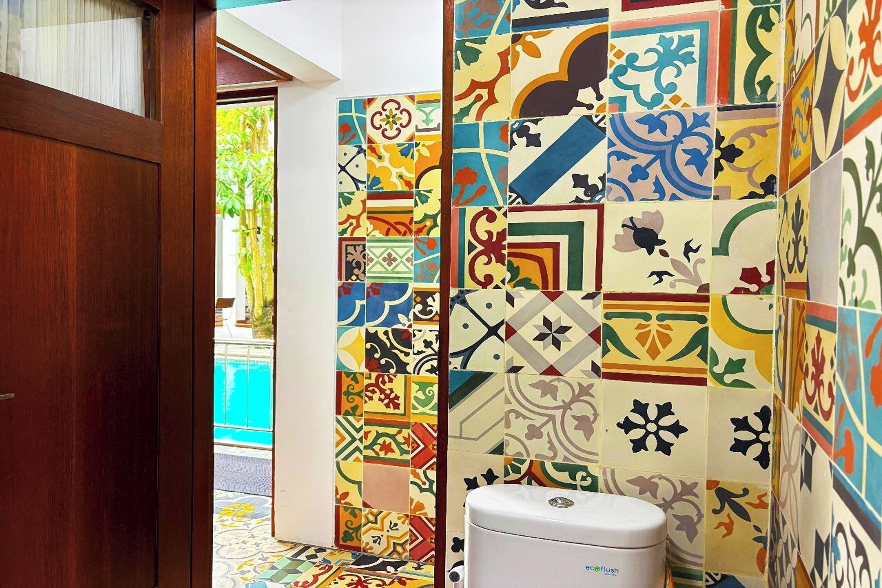 Une salle de bain avec des carreaux colorés à motifs, des toilettes blanches et une vue sur une piscine à l’extérieur.