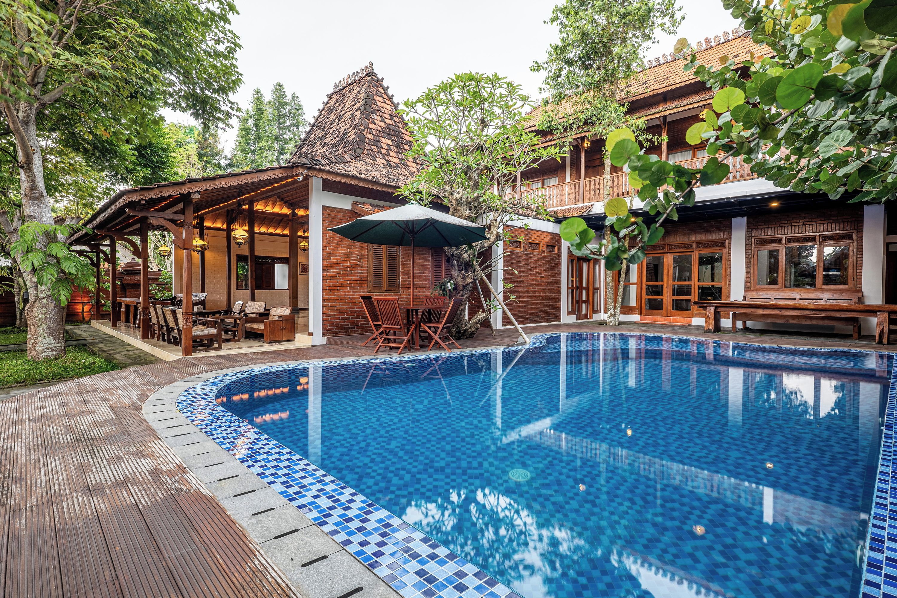 Una piscina con borde de azulejos, cubierta de madera y una zona de estar techada con sillas y mesas.