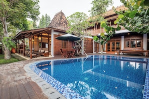 A swimming pool with a tiled edge, wooden deck, and a covered seating area with chairs and tables. - Joglo Wardani By Zuzu (Ngaglik)