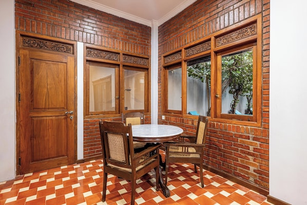 A corner room with a brick wall, wooden doors, a round table with chairs, and a window with a view of greenery. - Joglo Wardani By Zuzu (Ngaglik)