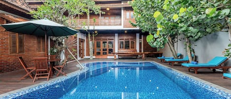A swimming pool with lounge chairs and a table, surrounded by a wooden deck and a brick building.