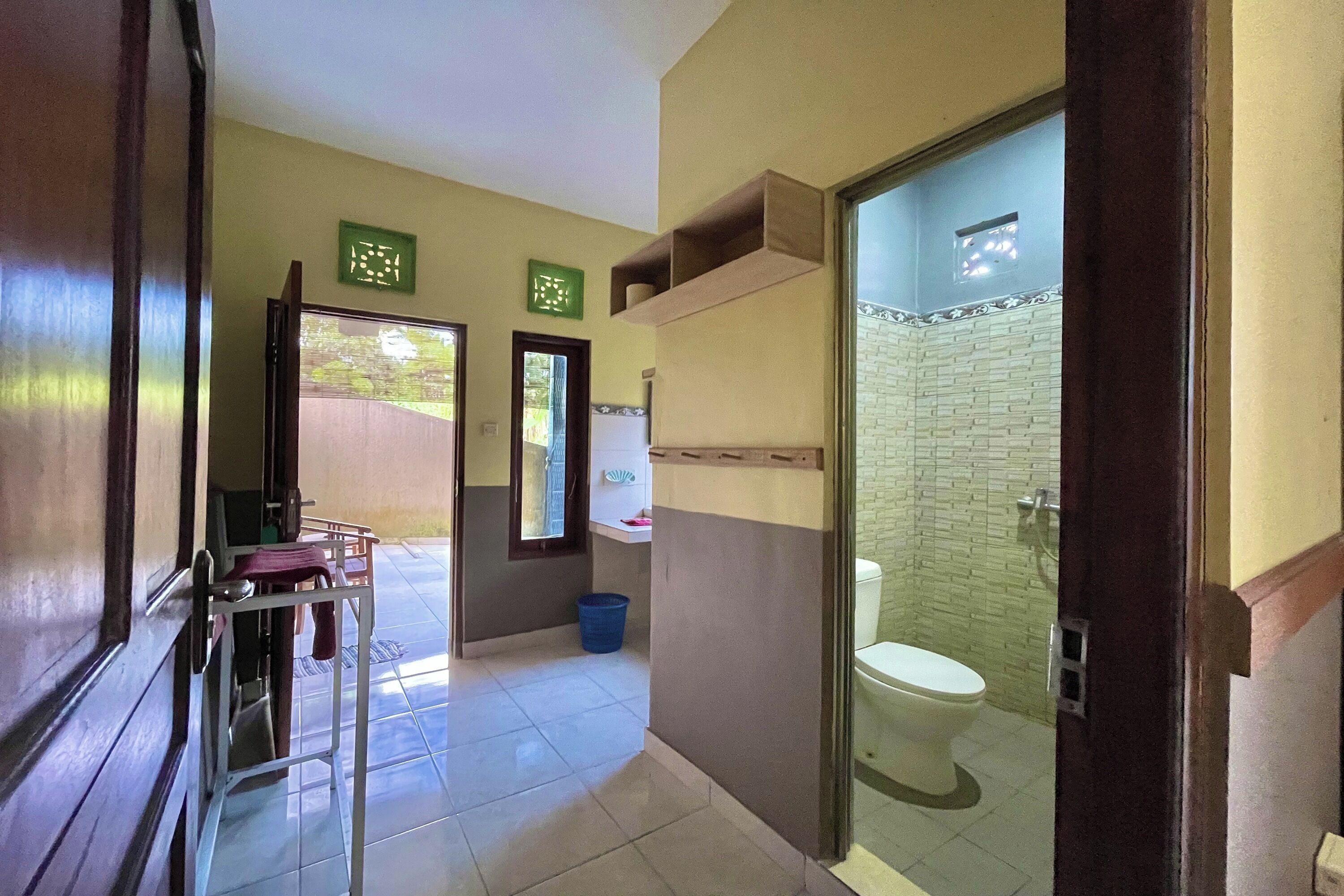 A bathroom with a toilet, a shower area, and a sink. There are two green signs on the wall, a blue bucket on the floor, and a door leading to another room.