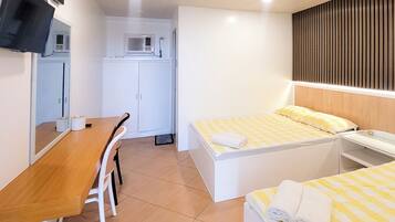Quarto de hotel com uma secretária de madeira, duas camas com lençóis xadrez amarelos, um guarda-roupa branco, uma televisão de ecrã plano montada na parede e uma janela com unidade de ar condicionado.