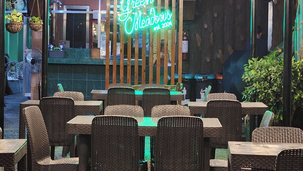 Un restaurante al aire libre con mobiliario de mimbre y un letrero de neón que dice "Green Meadows".