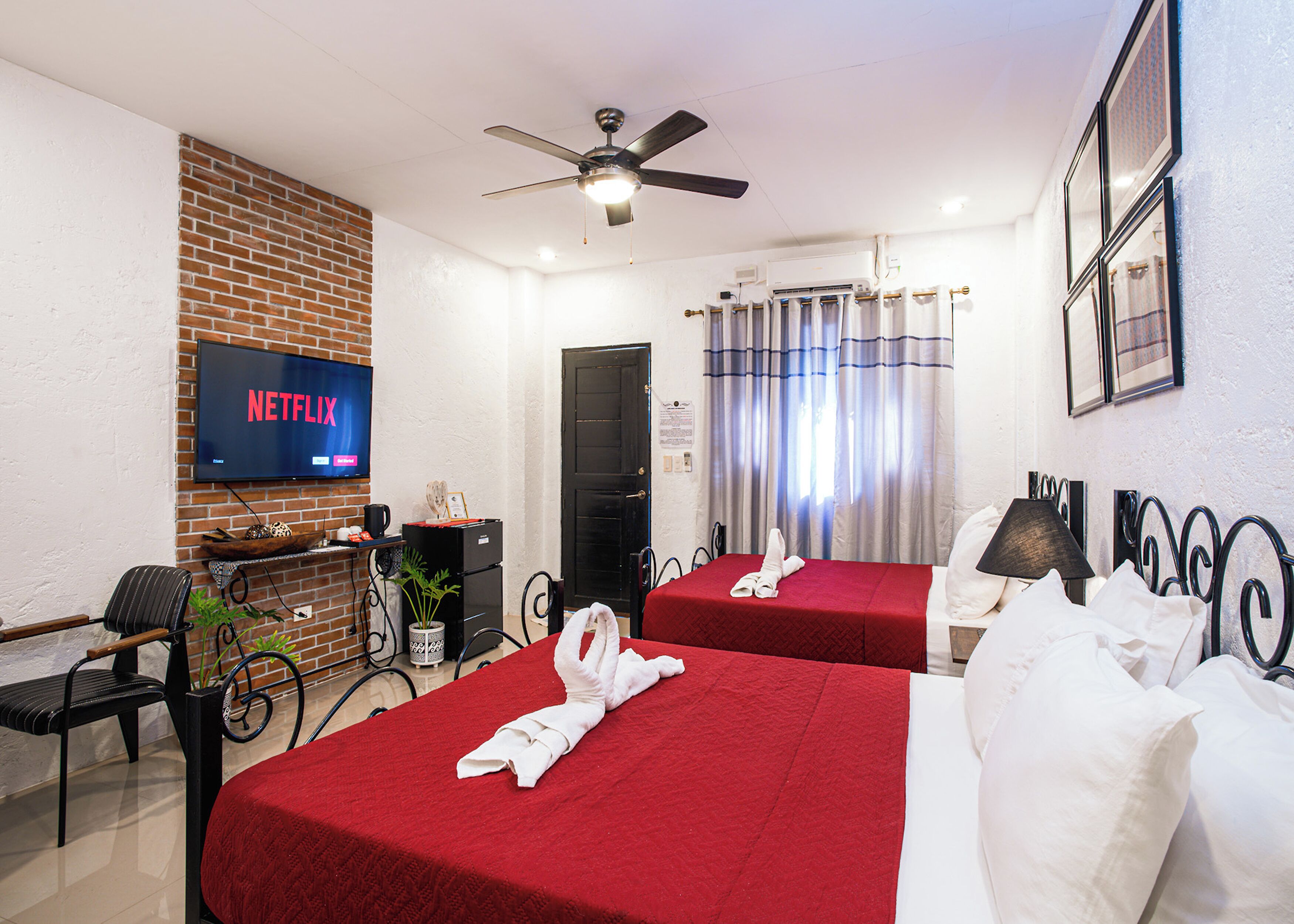 Une chambre d’hôtel avec deux lits, un téléviseur diffusant Netflix, un ventilateur de plafond et un mur en briques.